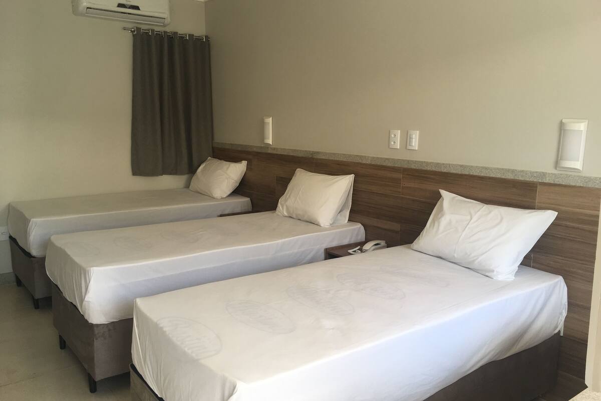 A hotel room with three single beds, a wooden headboard, an air conditioning unit, and a curtain.