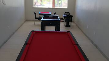 A pool table with a red felt surface, a foosball table, and two chairs in a room with a large window.