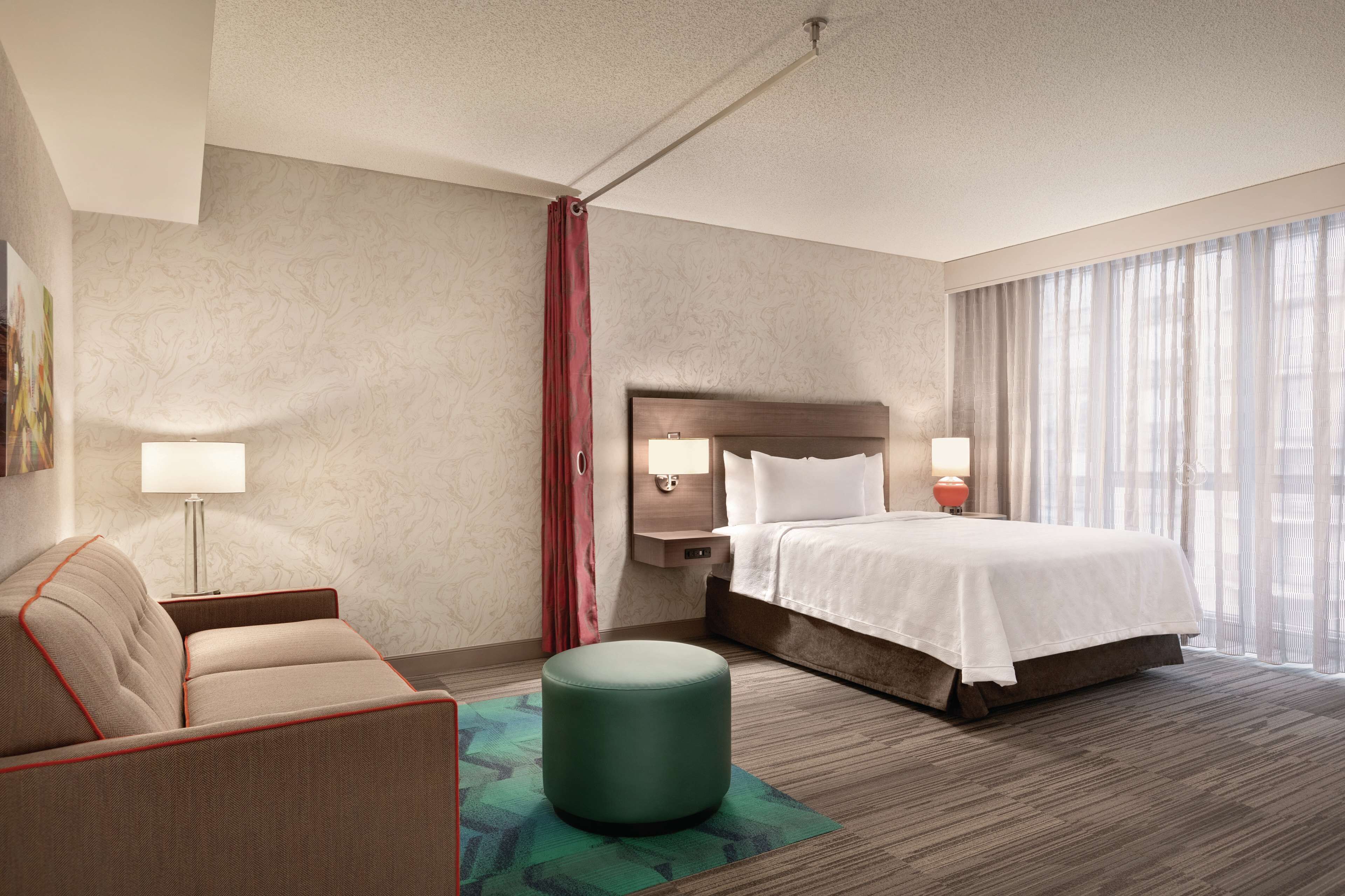 A hotel room with a bed, a sofa, a small ottoman, a nightstand, a lamp, and a window with curtains.