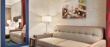 Habitación de hotel con sofá, mesita de noche con lámpara, un cuadro en la pared y un espejo que refleja la habitación.