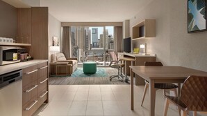 Ein modernes Hotelzimmer mit Küchenzeile, Essbereich, einer Sitzecke mit Blick auf die Stadt und einem großen Fenster.