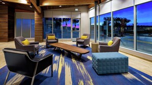Eine moderne Lobby mit Holzbalken an der Decke, bequemen Sitzgelegenheiten mit gelben Kissen, einem großen Holztisch und großen Fenstern mit Blick auf Palmen und einen Sonnenuntergang.