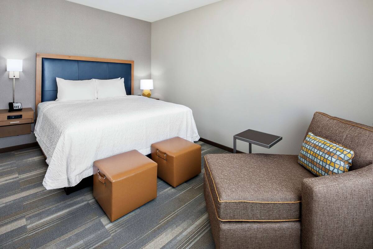 A hotel room with a large bed, two brown ottomans, a small table, and a chair with a patterned cushion.