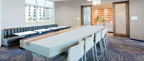 A hotel room with a long dining table, multiple chairs, a sofa, and a view of the outside.