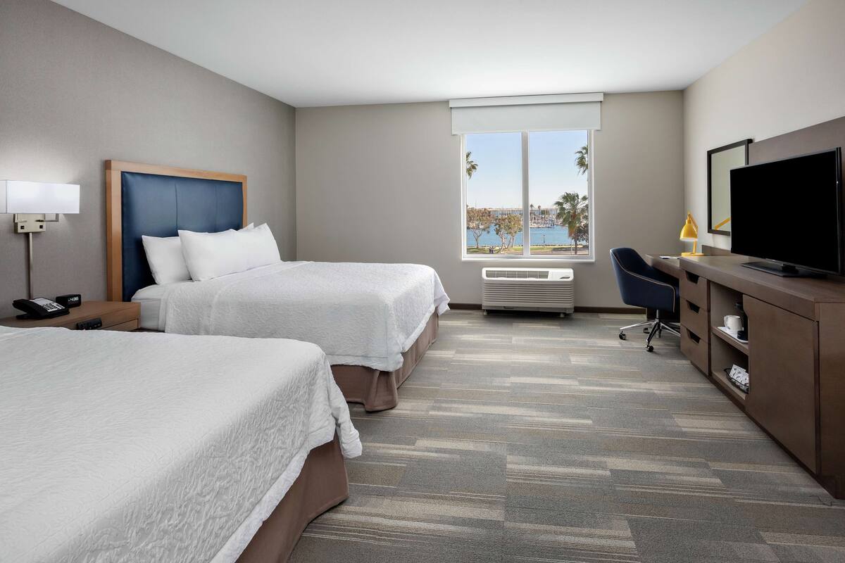 A hotel room with two beds, a desk, a TV, and a window with a view of palm trees and water.
