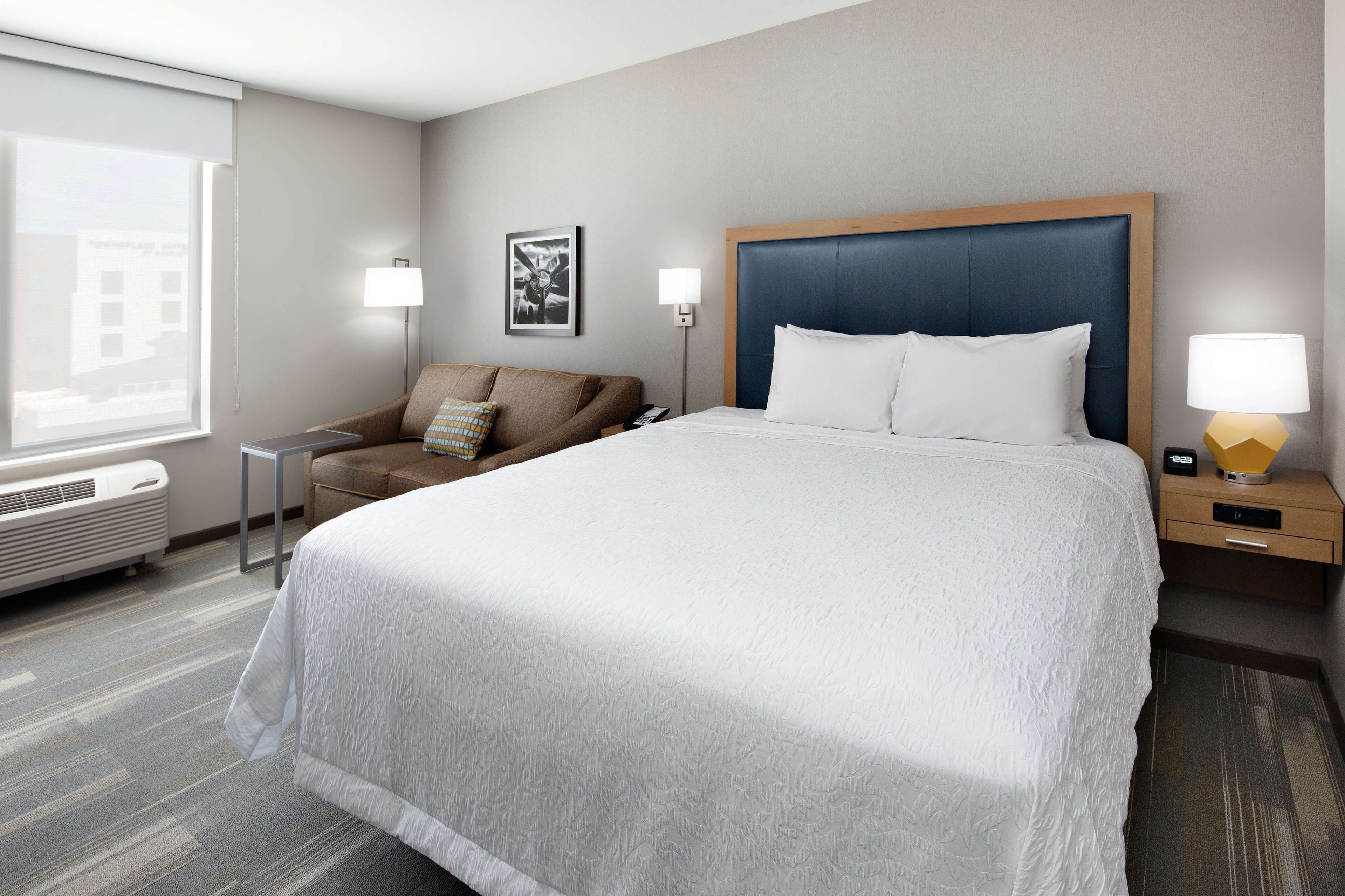 A hotel room with a large bed, a sofa, two bedside lamps, a heater, and a window with blinds.