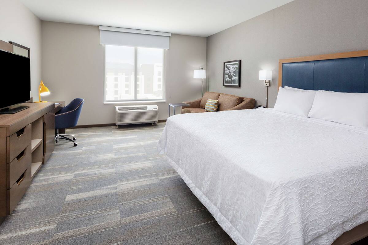 A hotel room with a large bed, a desk with a computer, a chair, a window with blinds, and a wall-mounted TV.
