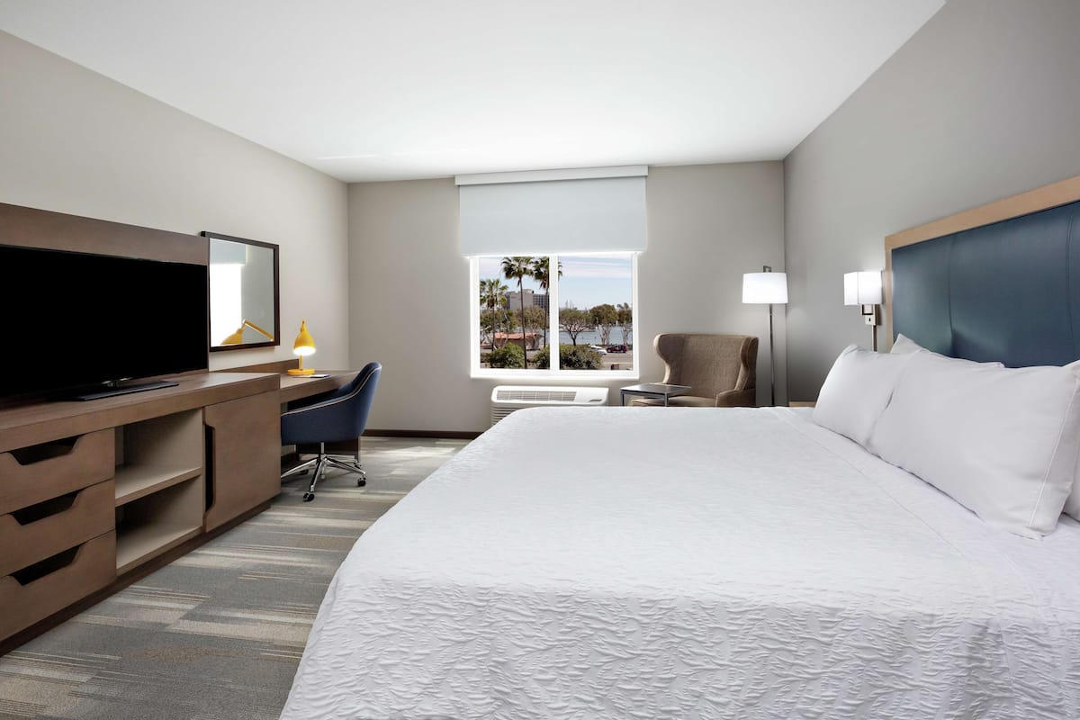 A hotel room with a large bed, a desk with a computer, a chair, a TV, and a window with a view of palm trees and buildings.