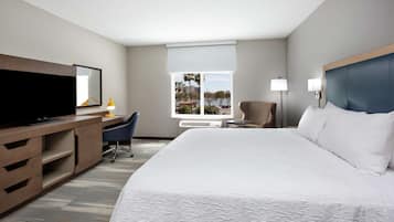 Quarto de hotel com uma cama grande, uma escrivaninha com um computador, uma cadeira, uma televisão e uma janela com vista para palmeiras e edifícios.