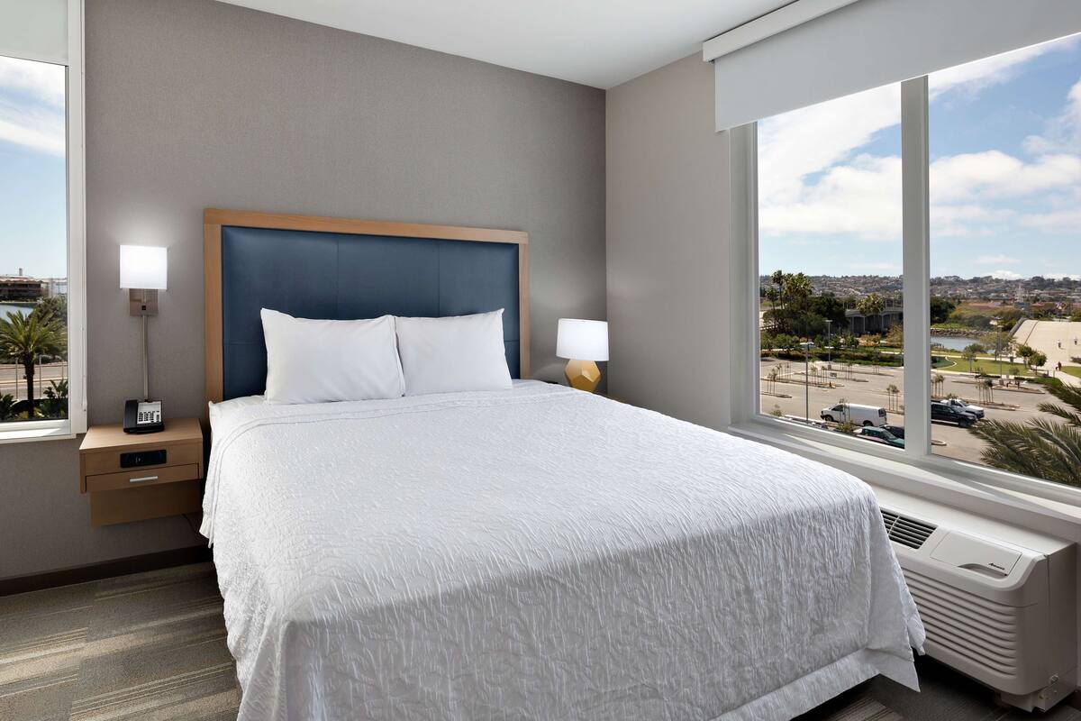 A hotel room with a large bed, a nightstand with a lamp, a window with a city view, and a wall-mounted heater.
