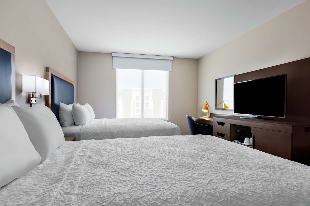 A hotel room with two beds, a desk, a television, and a window with a view of buildings.