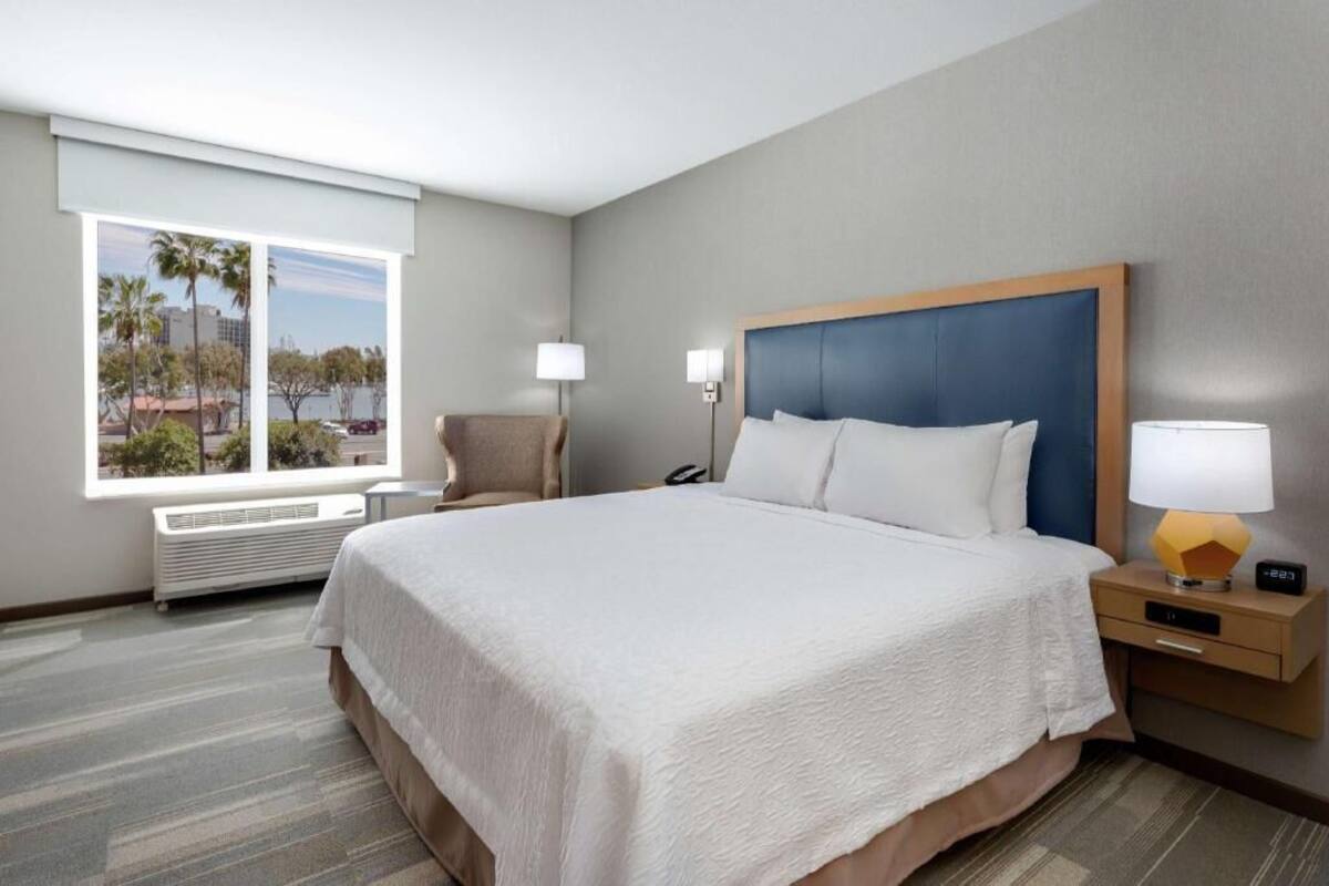 A hotel room with a large bed, a chair, a nightstand with a lamp, a phone, and a window with a view of palm trees and buildings.