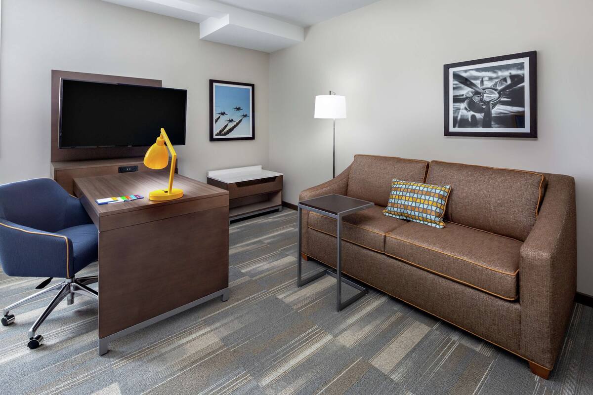 A hotel room with a desk, a blue office chair, a brown sofa with a patterned pillow, a flat-screen TV, a framed picture of planes, a floor lamp, and a small table.