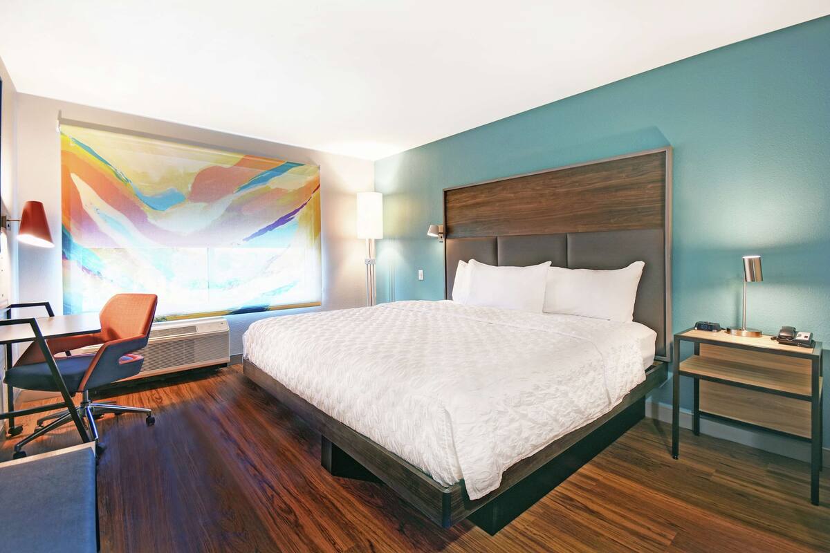 A modern hotel room with a large bed, a desk, and a colorful abstract painting on the wall.