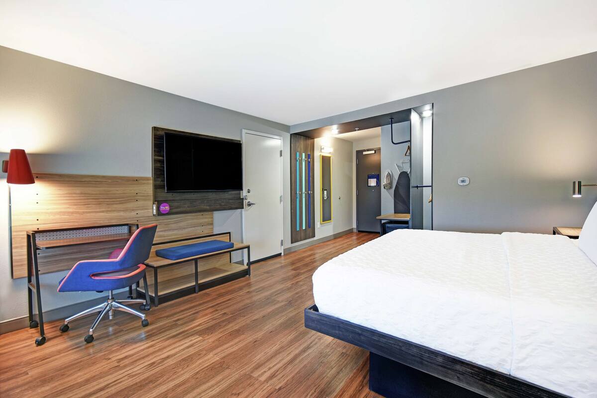 A hotel room with a bed, a desk with a chair, a TV, and a bathroom visible through an open door.