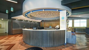 A modern hotel reception area with a curved counter, a digital display, and a view of the lobby. - Tru by Hilton Grand Rapids Airport (Grand Rapids)