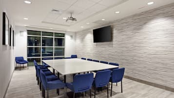 Uma sala de conferências com uma mesa grande, cadeiras azuis, uma TV fixada na parede e um papel de parede estampado.