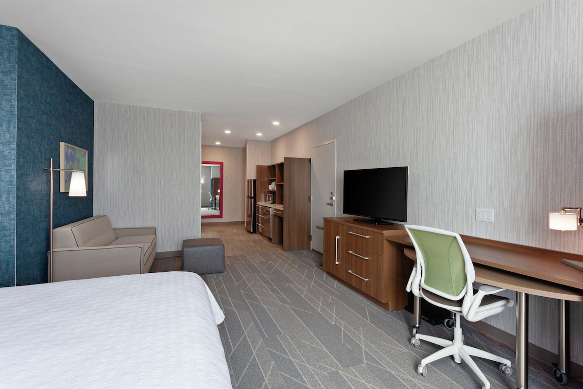 A hotel room with a bed, a desk with a chair, a television, and a kitchenette.