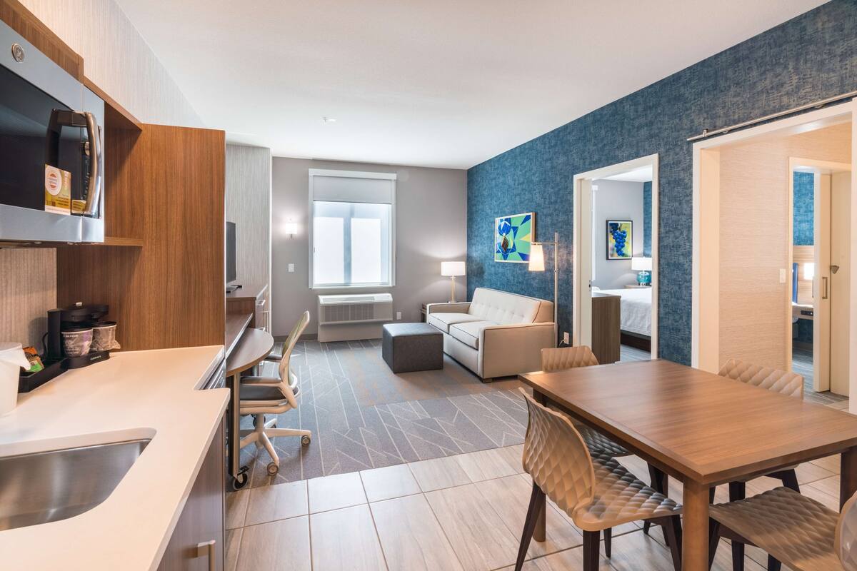 A modern kitchen with a built-in microwave, a coffee machine, and a sink. There is a dining area with a wooden table and chairs, and a living area with a sofa and a coffee table.