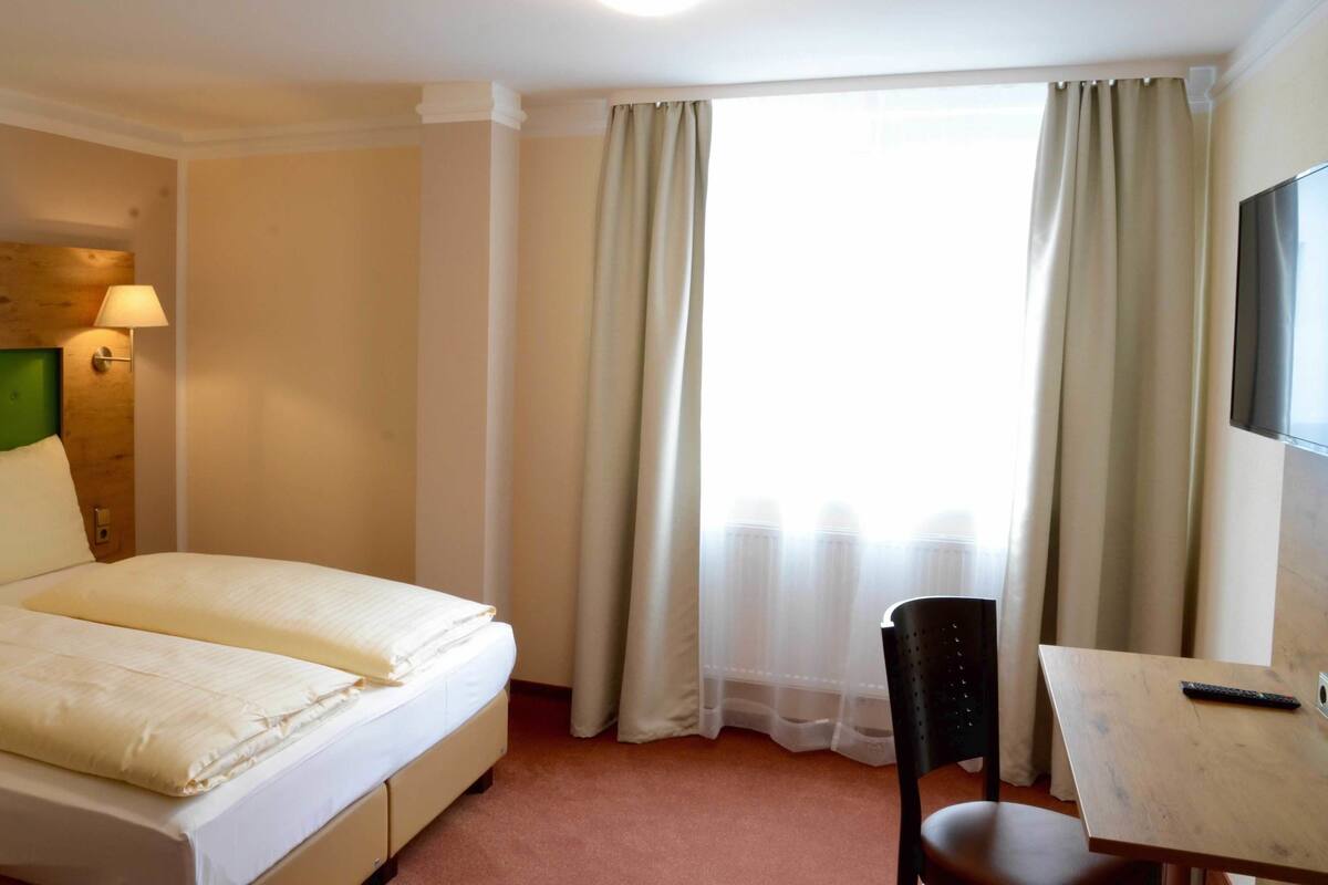 Ein Hotelzimmer mit Bett, Schreibtisch, Stuhl, Lampe und einem fenster mit Vorhängen.