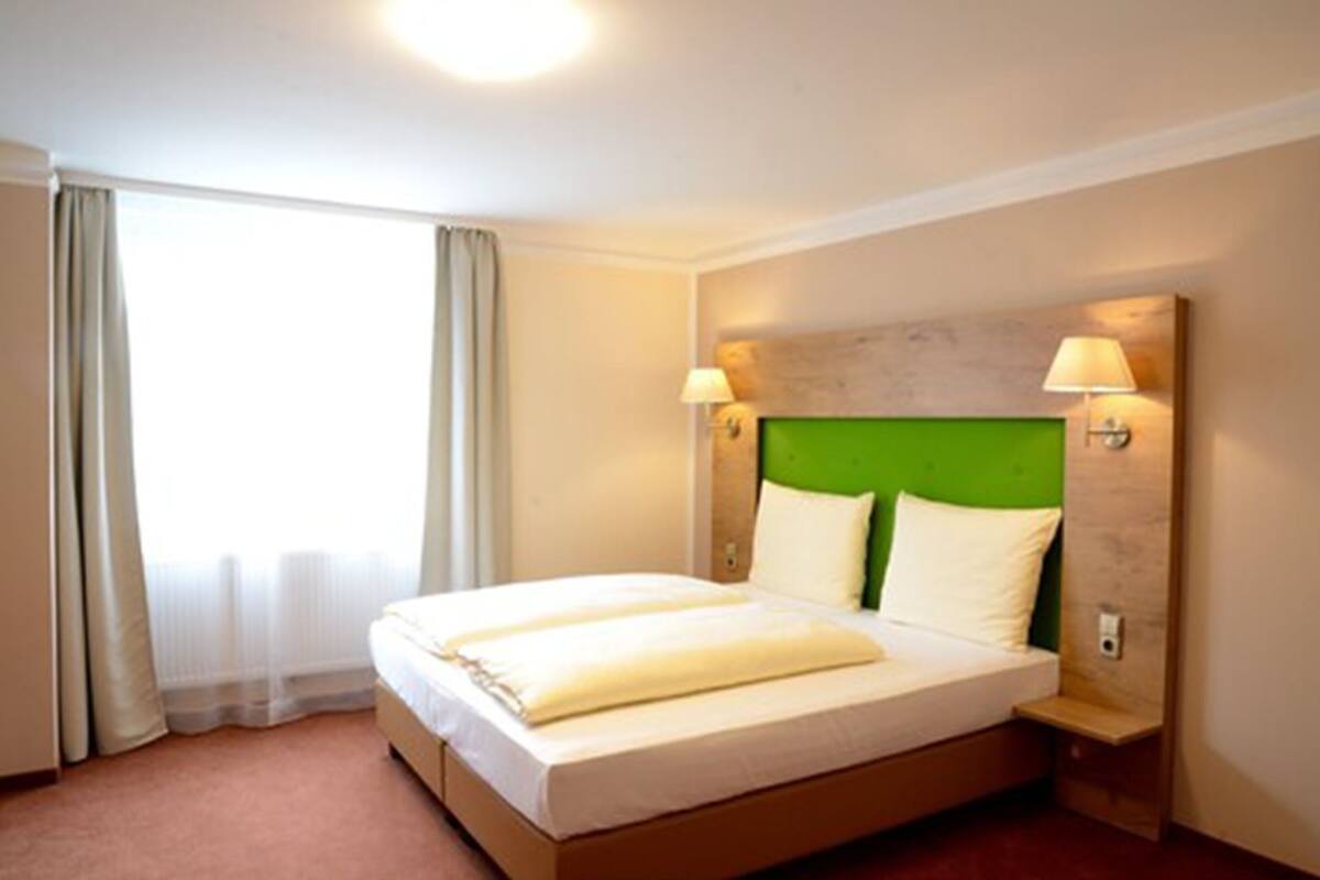 Ein Hotelzimmer mit Bett, grünem Kopfteil, zwei Nachttischlampen und einem Fenster mit Vorhängen.