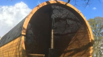 Eine zylindrische Holzkonstruktion mit einer zentralen Säule, möglicherweise ein Denkmal oder eine Skulptur, auf einer gepflasterten Fläche inmitten von Gras und Bäumen.