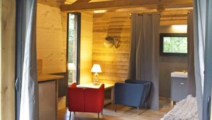 A cozy cabin interior with a wooden interior, a small desk with a lamp, a blue chair, a red chair, and a bed with a blanket. - Ecolodges de Loire & Spa (Gennes-Val-de-Loire)