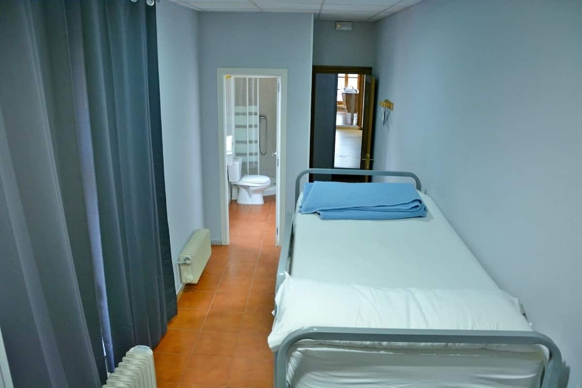 A narrow hallway leading to a bathroom with a toilet and a shower. There are two beds with white linens and a blue blanket.