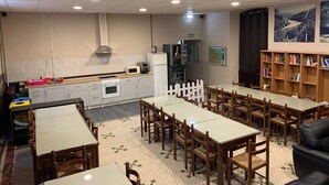 A dining area with multiple tables and chairs, a kitchen area with appliances, and a bookshelf with books. - Albergue La Estación - Hostel (Llanes)
