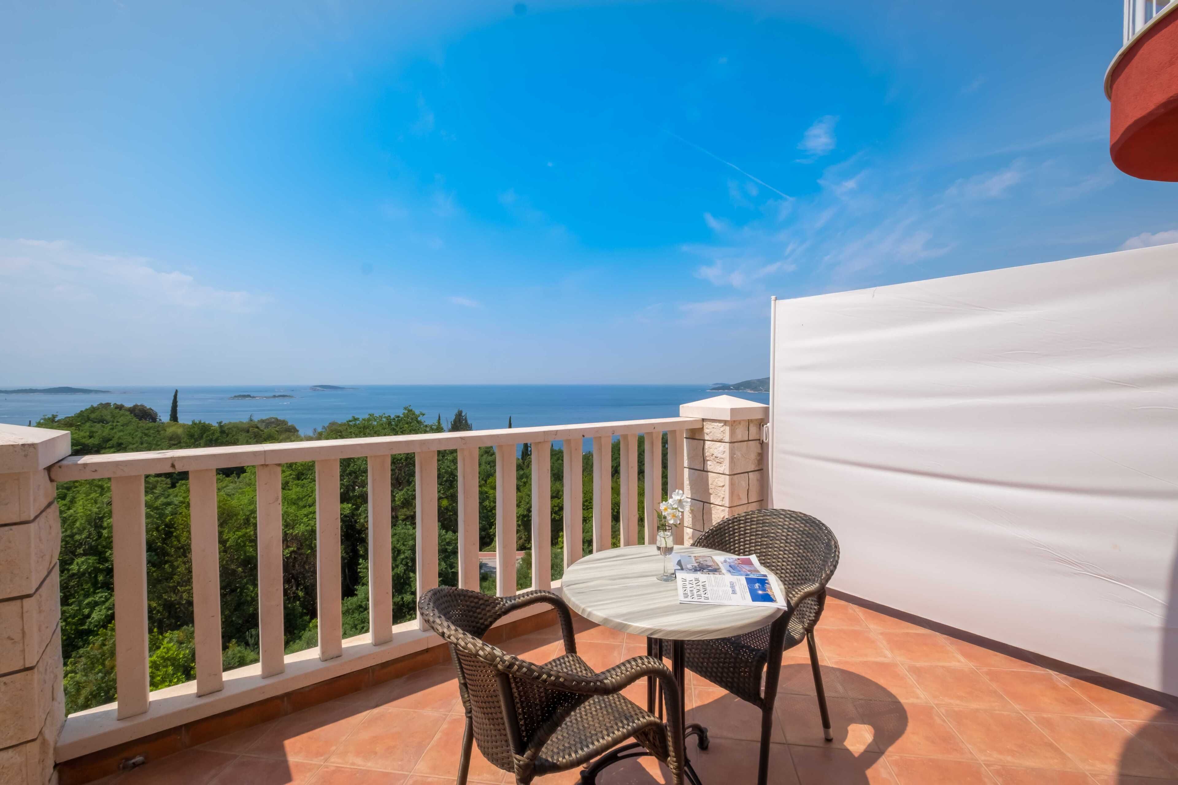 Varanda com duas cadeiras de vime e uma mesa pequena, com vista panorâmica para o mar e vegetação.