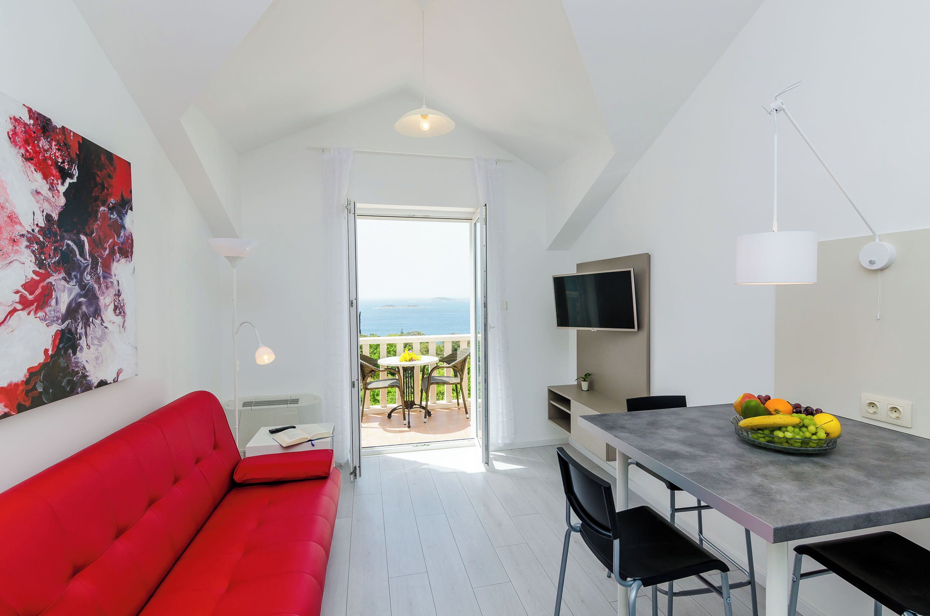 Una habitación moderna con un sofá rojo, una mesa de comedor con fruta, un televisor y un balcón con vistas.