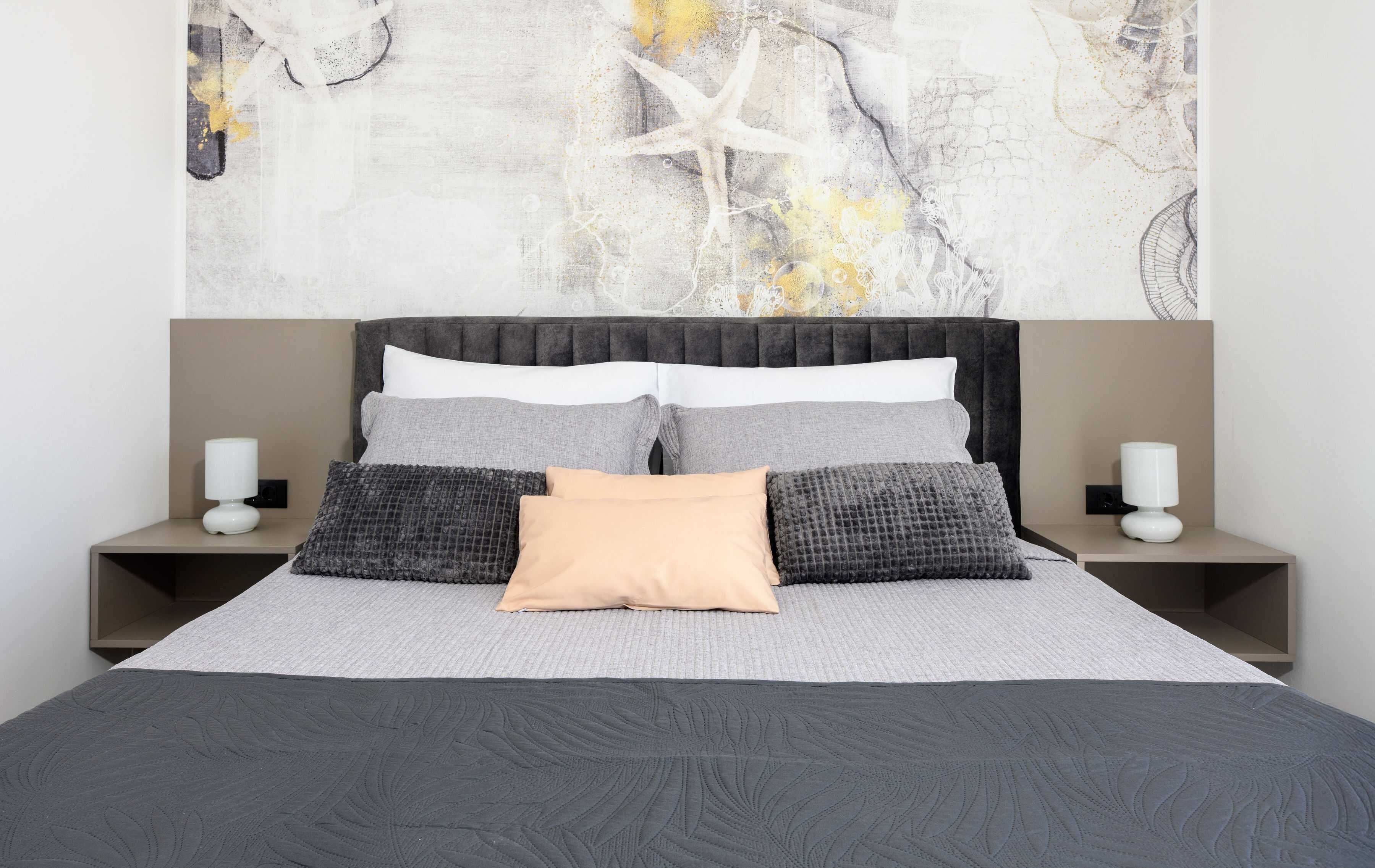 Una cama bien hecha con un edredón gris y blanco estampado, una almohada beige central y dos almohadas grises estampadas. Hay dos mesitas de noche con lámparas blancas.