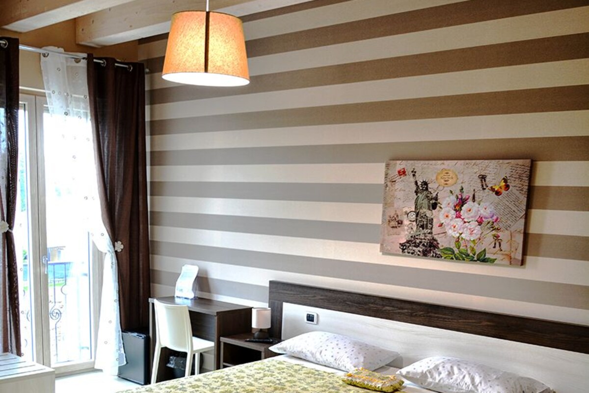 A hotel room with a bed, a desk, a chair, a lamp, and a painting on the wall.