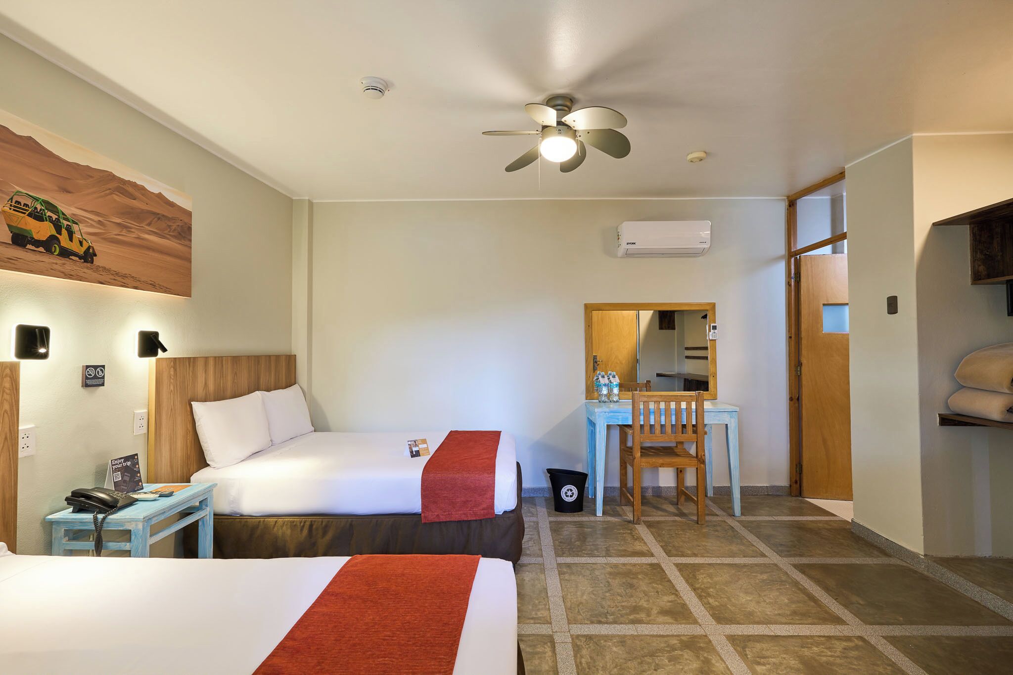 A hotel room with two beds, a desk, a chair, a ceiling fan, an air conditioning unit, and a small trash bin.