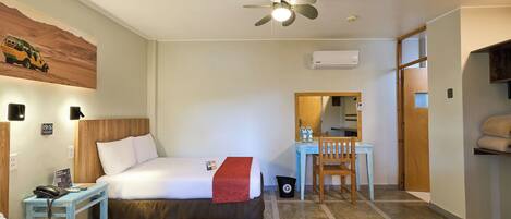 A hotel room with two beds, a desk, a chair, a ceiling fan, an air conditioning unit, and a small trash bin.