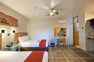 Habitación de hotel con dos camas, un escritorio, una silla, un ventilador de techo, un equipo de aire acondicionado y un pequeño cubo de basura.