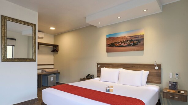 A hotel room with a large bed, a bedside table, a nightstand, a mirror, a painting of a desert landscape, and a red throw blanket. - Casa Andina Standard Ica   (Ica)