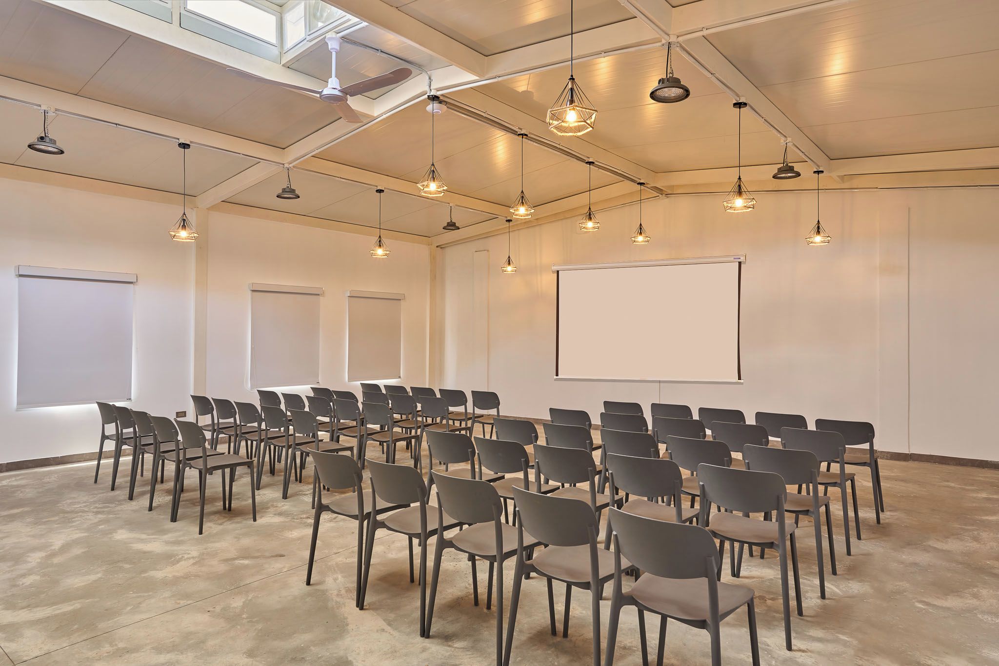 Un salón de conferencias con filas de sillas, una pantalla de proyector e iluminación colgante.