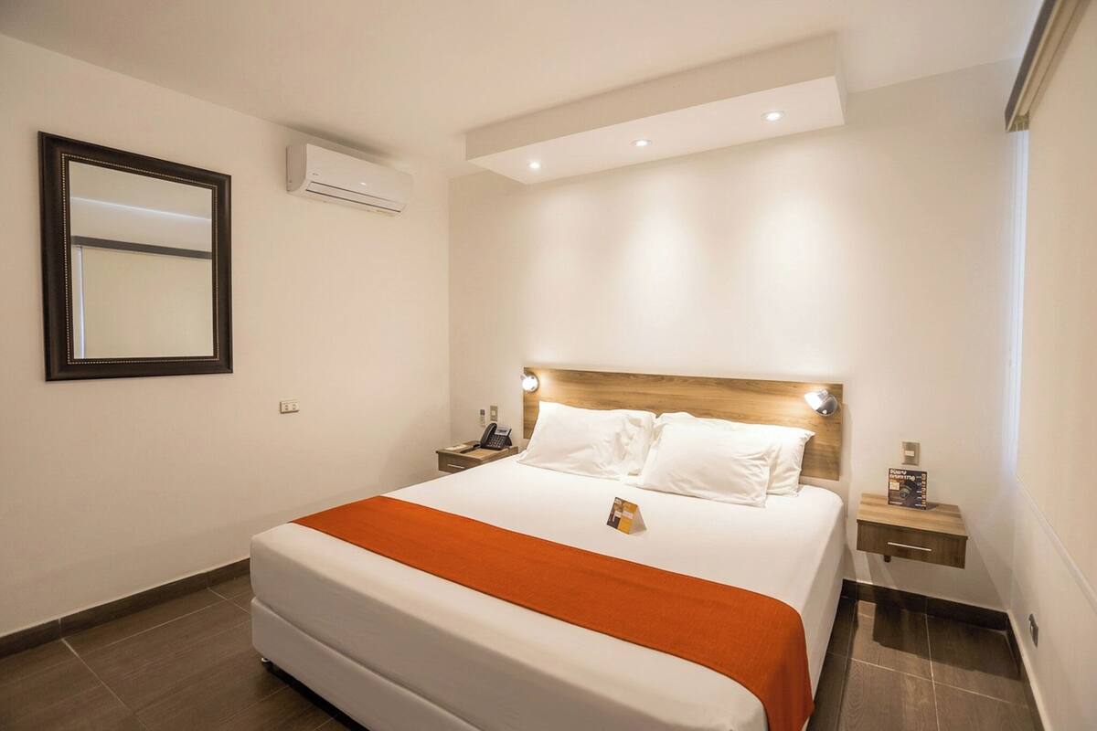 A hotel room with a large bed, a bedside table, an air conditioning unit, and a wall-mounted mirror.
