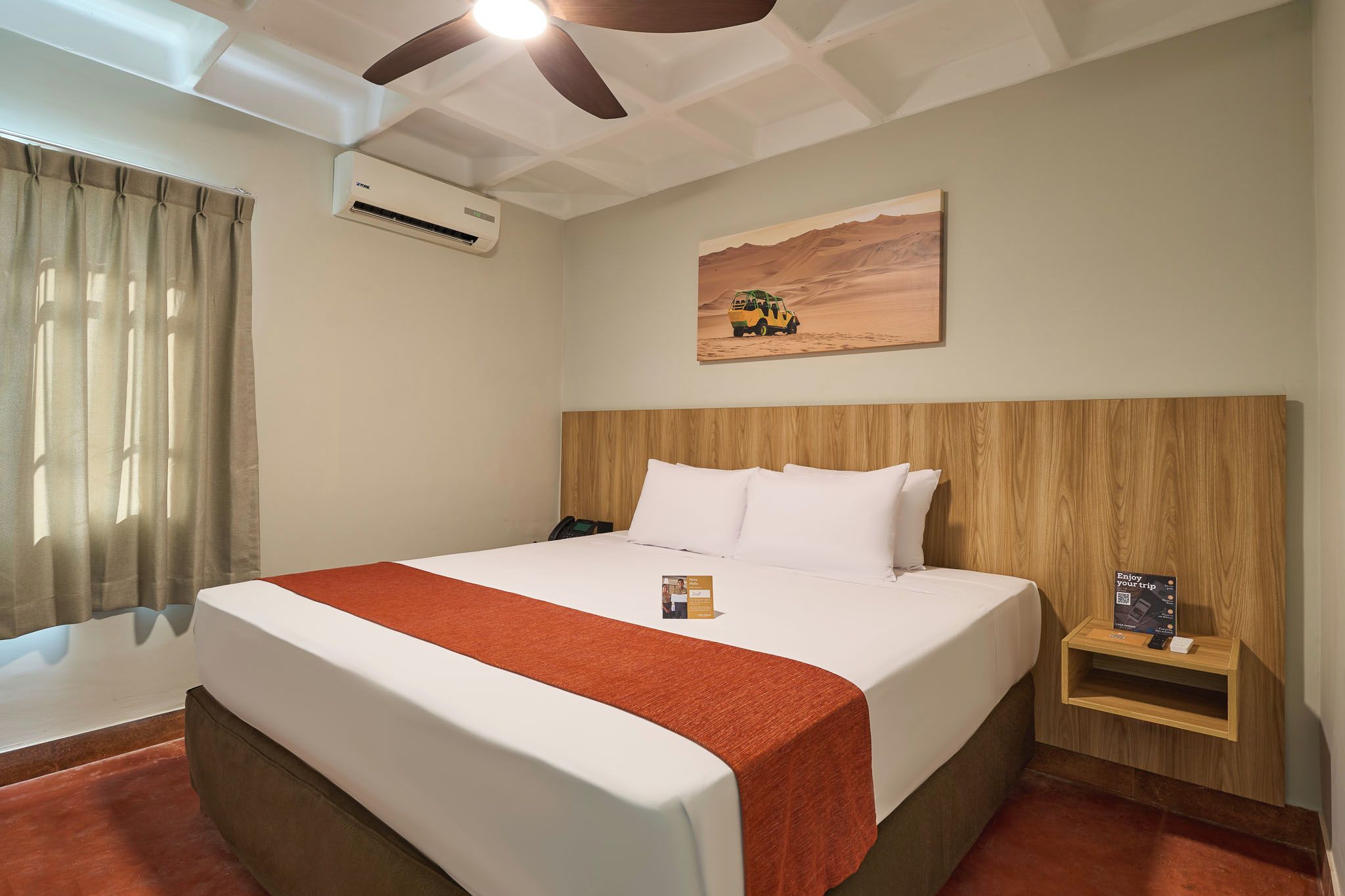 A hotel room with a bed, a nightstand, a wall-mounted air conditioner, a painting of a desert landscape, and a ceiling fan.