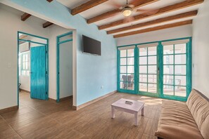 A spacious living area with a wooden floor, a beige sofa, a small table, and a ceiling fan. There are two doors with teal frames leading to other rooms, and a large window with a view. - Casa Andina Standard Ica   (Ica)