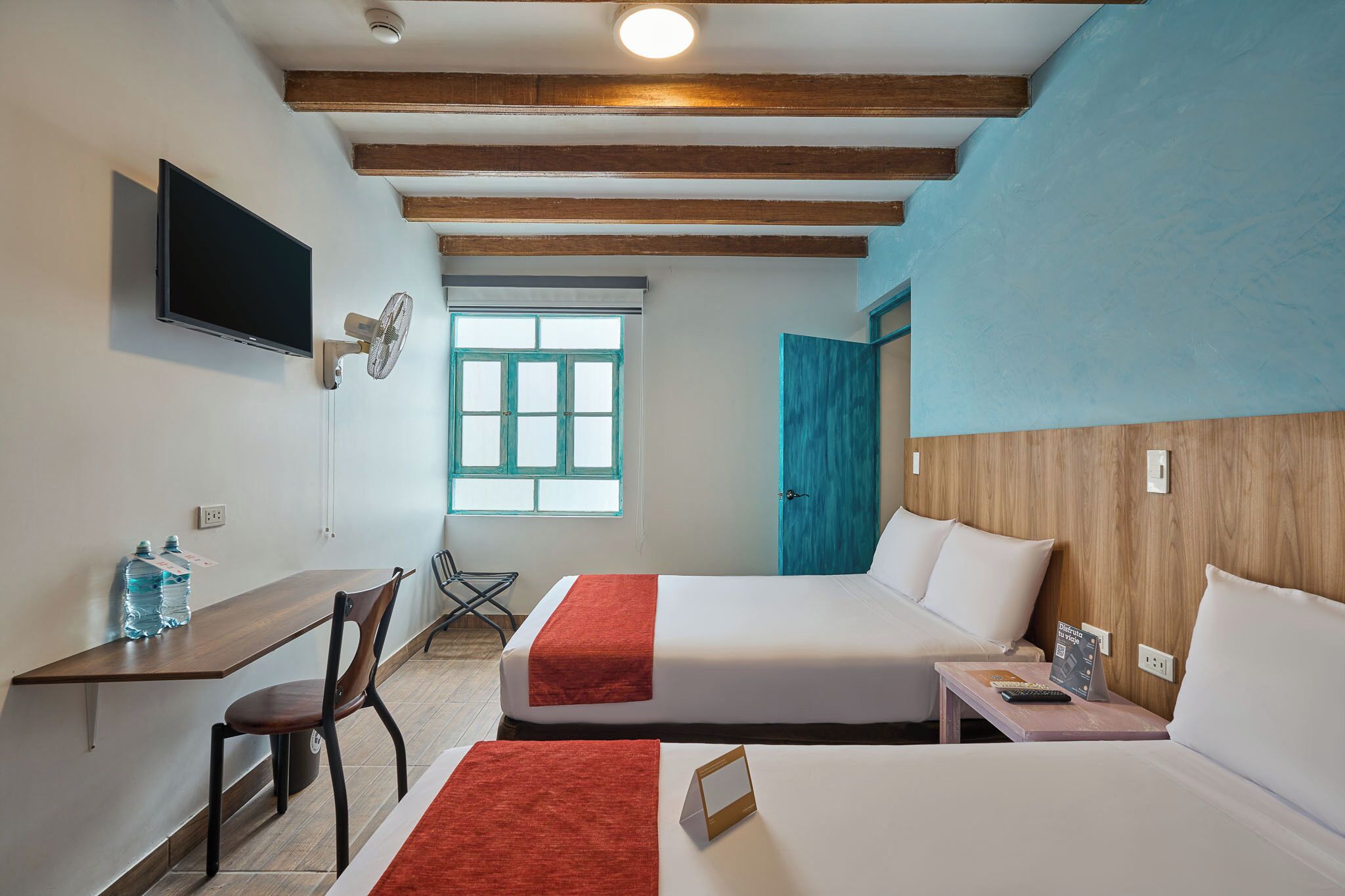 A hotel room with two beds, a desk, a chair, a TV, a window, and a door.