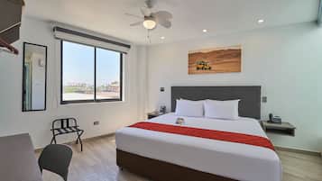 A hotel room with a large bed, a desk, a chair, a window with a city view, a ceiling fan, and a painting on the wall.