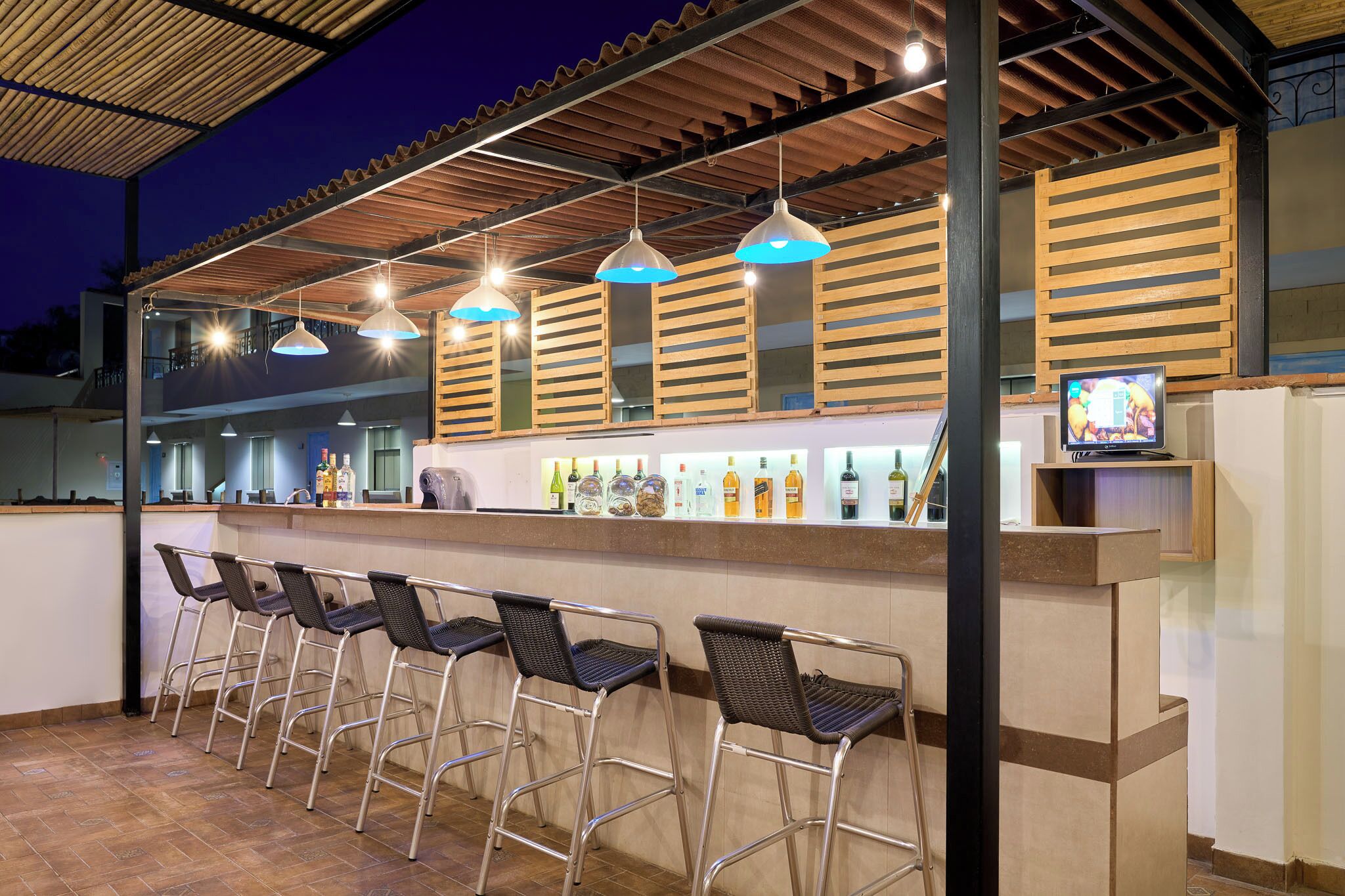 An outdoor bar area with metal chairs, a counter with bottles, and hanging pendant lights.