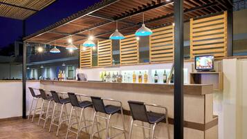 An outdoor bar area with metal chairs, a counter with bottles, and hanging pendant lights.