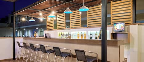 An outdoor bar area with metal chairs, a counter with bottles, and hanging pendant lights.