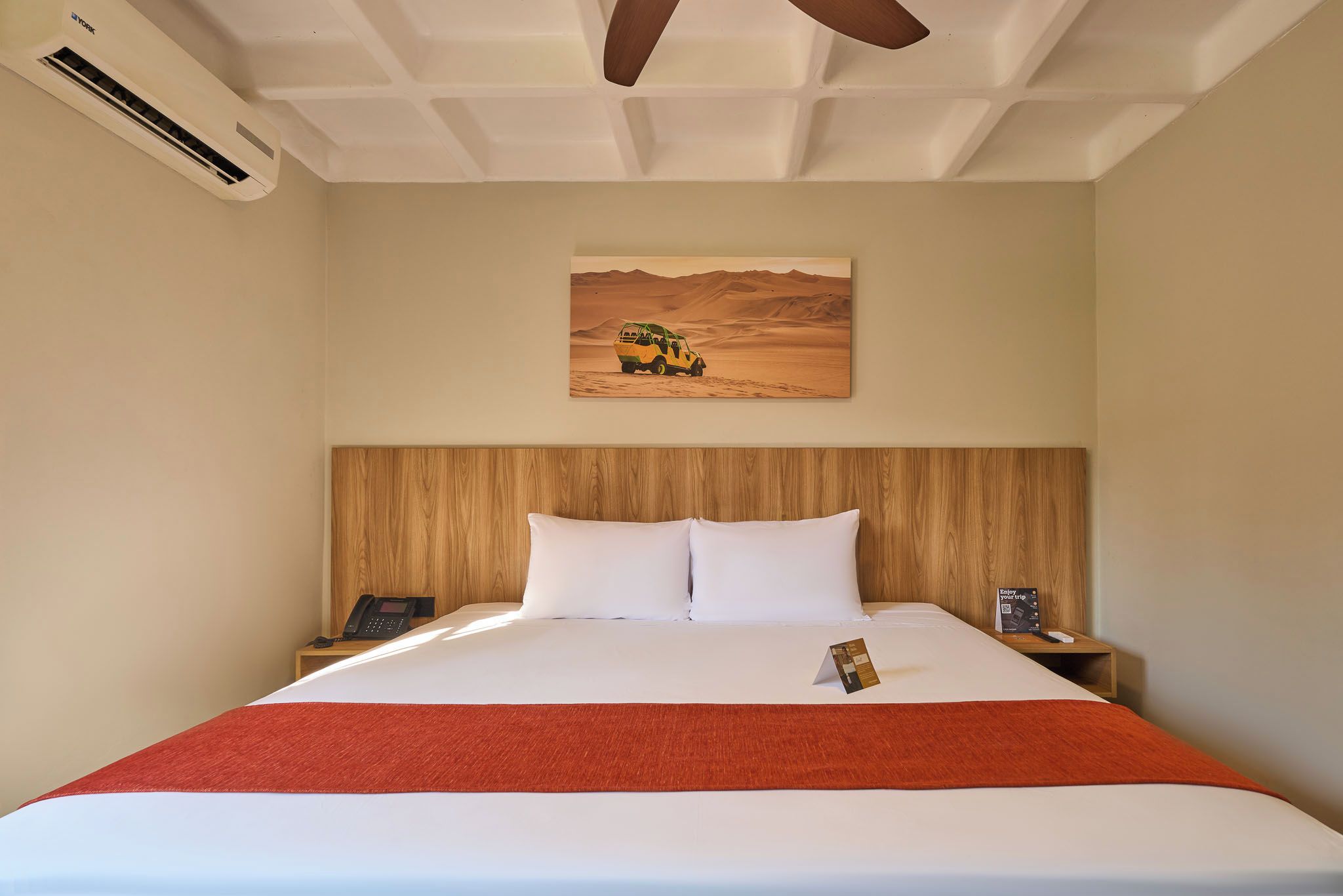 A hotel room with a bed, a wooden headboard, a painting of a desert vehicle, a telephone, and a brochure on the nightstand.