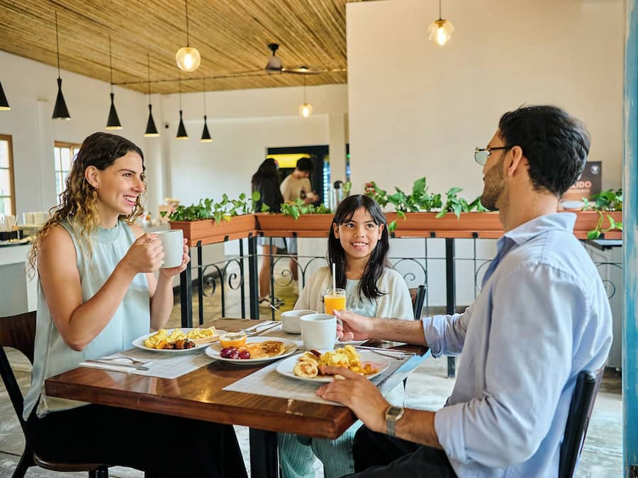 Un groupe de personnes est assis à une table en bois dans un café, sous des lampes suspendues, et des plantes en pot décorent l’endroit.