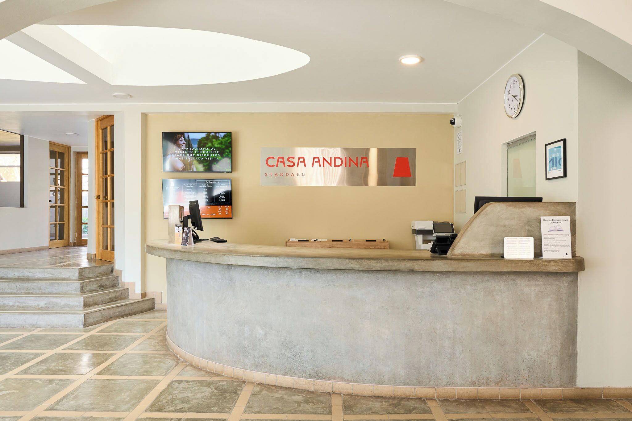 A reception desk with a clock, a TV screen displaying a program, and a sign that reads 'CASA ANDINA STANDARD'.