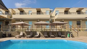 Una piscina con sillones de descanso y sombrillas, rodeada por un edificio con balcones y un muro de piedra.